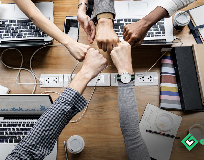 NewBossCommunications. A birds eye photo of a desk with 5 people fistbumping over laptops.